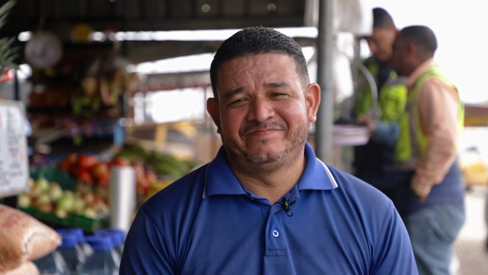 Un hombre con camisa azul sonríe frente a un puesto de frutas y verduras en un mercado